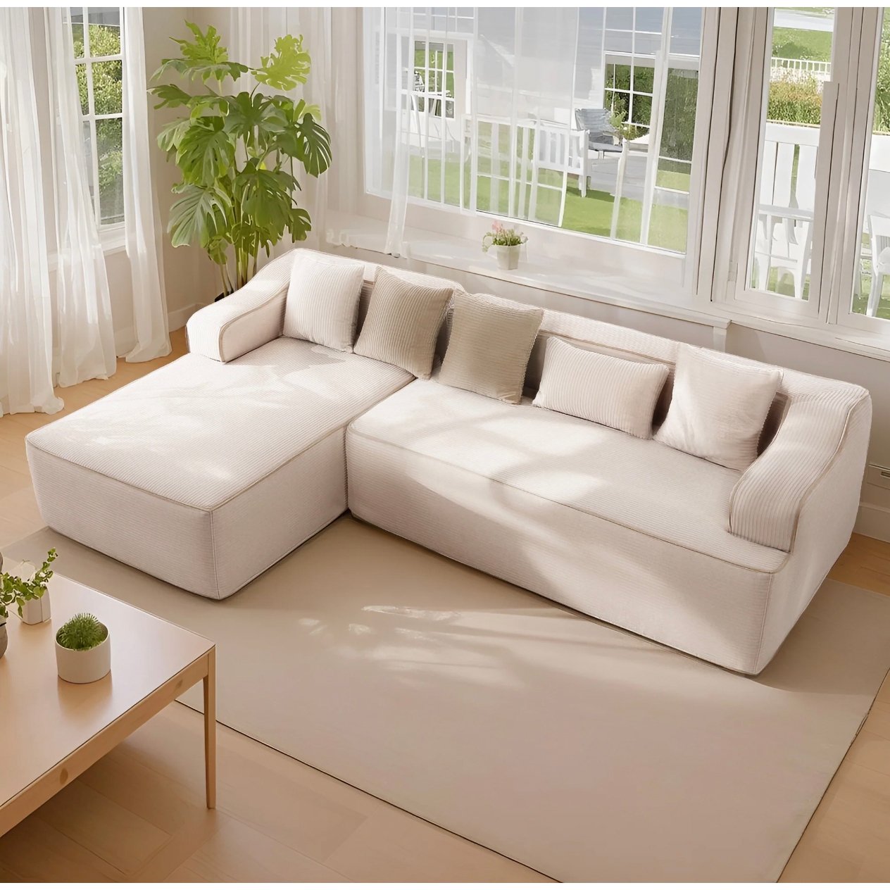 A cream-colored L-shaped sofa with a chaise lounge features matching cushions and rounded arms. It's placed on a light rug near a coffee table, with a potted plant nearby and large windows in the background.