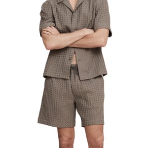 A man is wearing a textured short-sleeve button-up shirt and shorts set in a muted brown color.