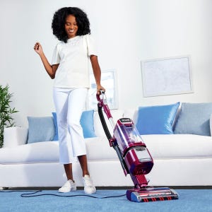 A woman using an upright Shark vacuum cleaner on a carpeted floor indoors.