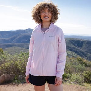 A woman stands outdoors wearing a light pink Columbia jacket and black shorts.