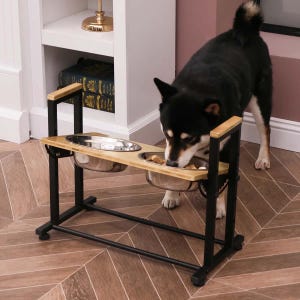 A dog is eating from a dual elevated pet feeder with stainless steel bowls on a wooden and metal stand.