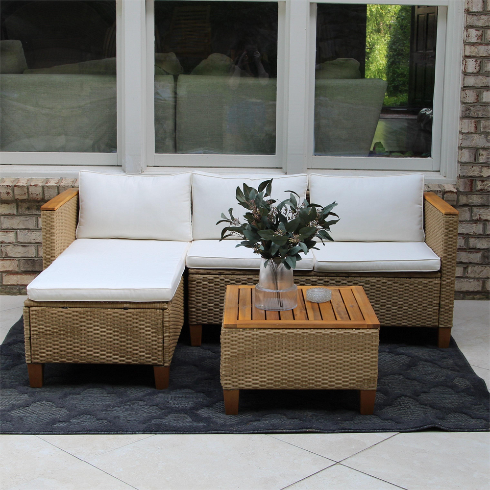 Outdoor wicker sectional sofa with white cushions, featuring a matching wicker coffee table with a wooden top and a vase of greenery on it, set on a dark patterned rug against a brick wall.