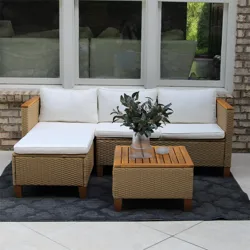 Outdoor wicker sectional sofa with white cushions, featuring a matching wicker coffee table with a wooden top and a vase of greenery on it, set on a dark patterned rug against a brick wall.
