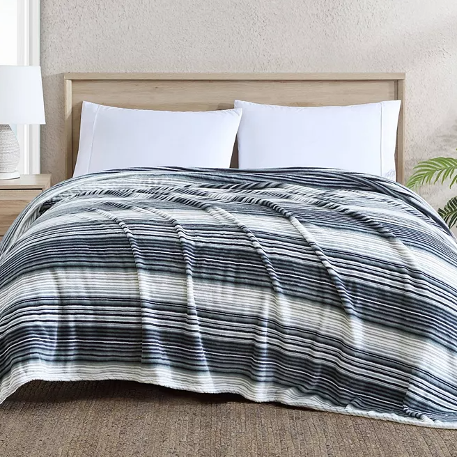 A striped comforter with varying shades of blue on a bed with white pillows and a wooden headboard.