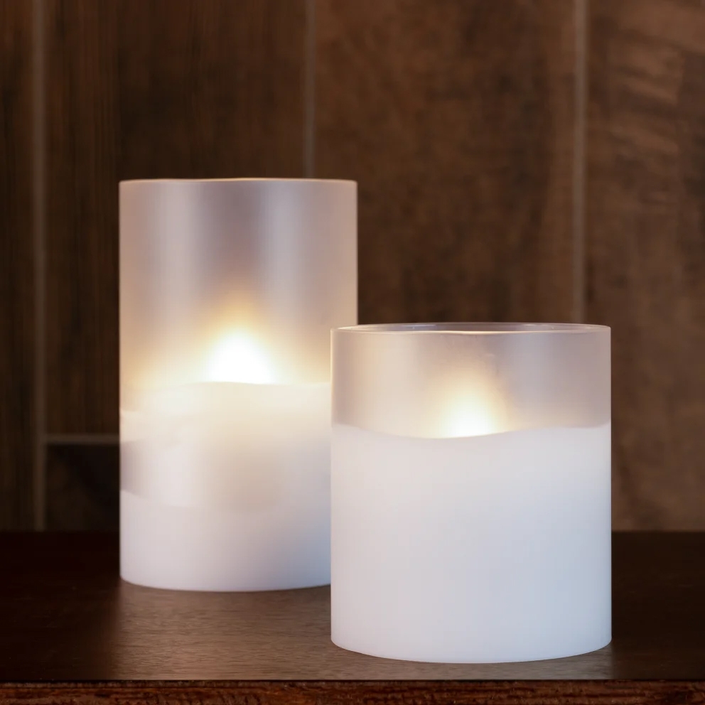 Two frosted LED candles of different heights emit a warm glow on a wooden surface.