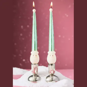 Two green-striped taper candles with cute snowman faces near the base, set in silver holders on a snowy surface, against a festive pink background.