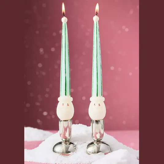 Two green-striped taper candles with cute snowman faces near the base, set in silver holders on a snowy surface, against a festive pink background.
