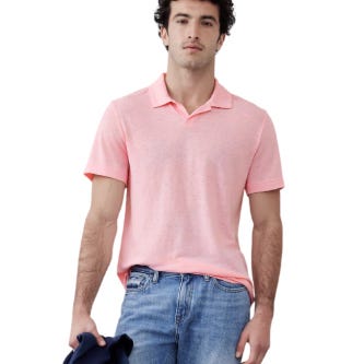 A man is wearing a light pink polo shirt paired with blue jeans, holding a dark jacket in his left hand.