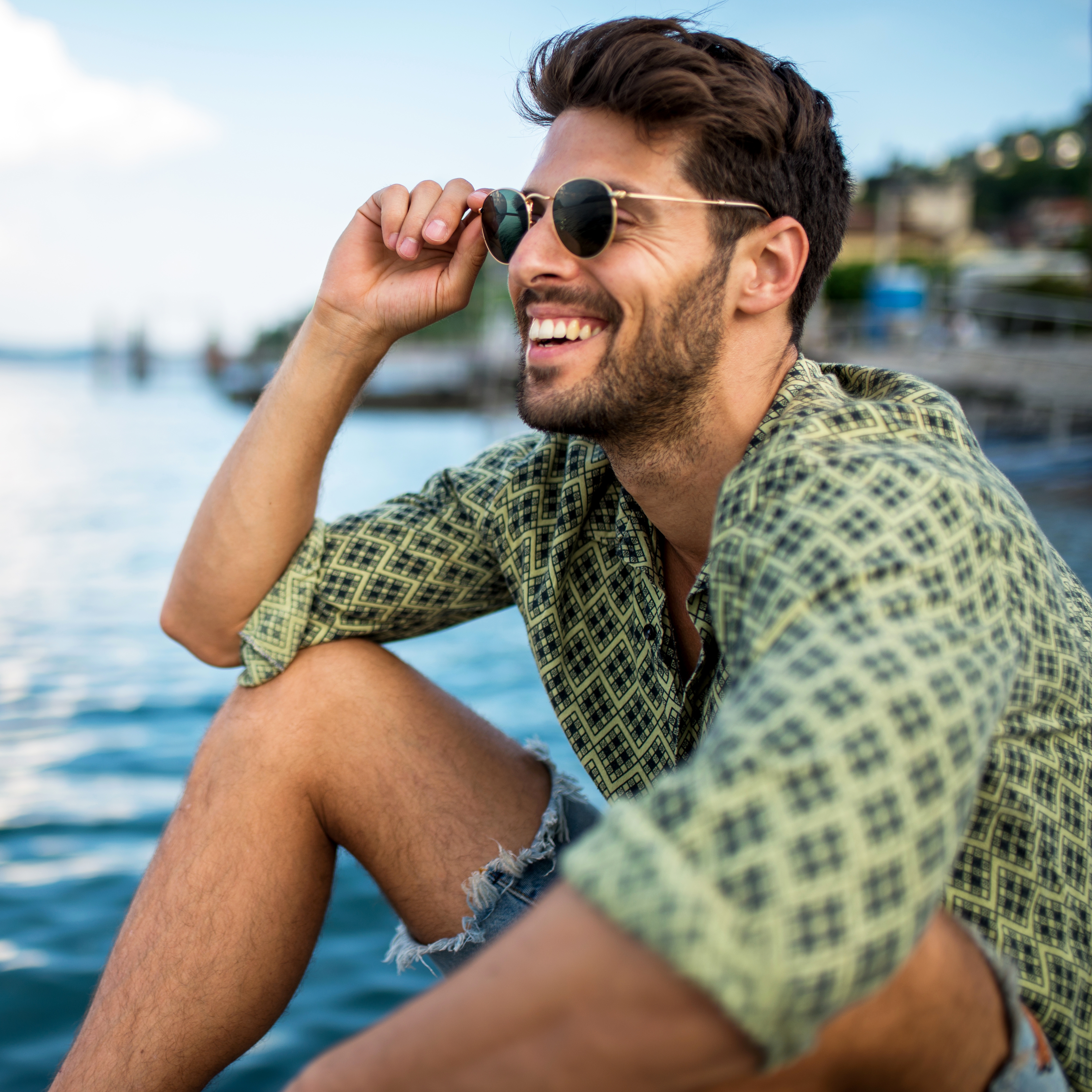 A man wearing round sunglasses, a patterned shirt, and distressed denim shorts.