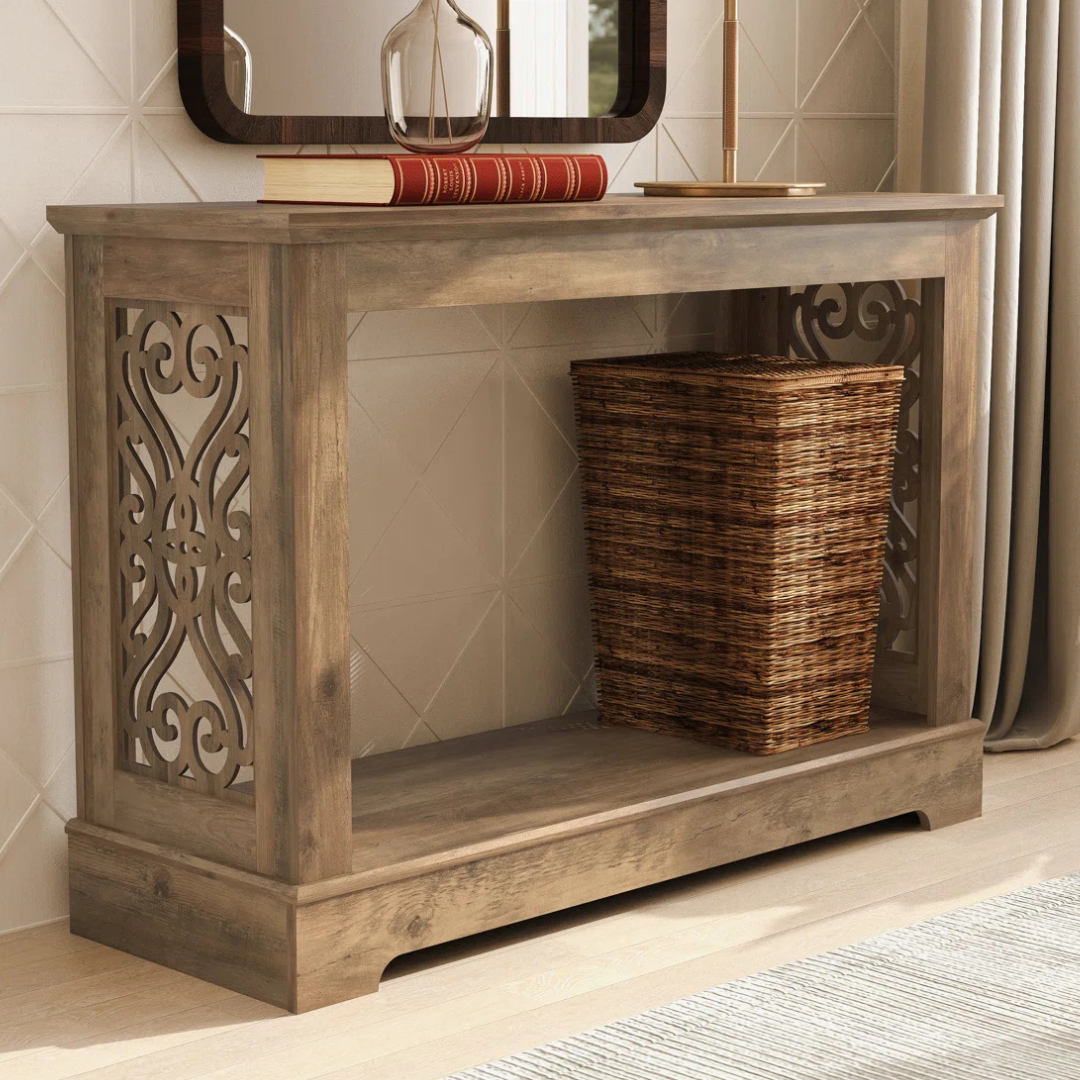 A wooden console table features intricate side cutouts, a flat top with books, a glass bottle, a lamp, and a wicker basket on the lower shelf.