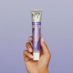 A hand is holding a tube of No7 Pure Retinol Post Retinol Soother, featuring a lavender to white gradient color scheme with text indicating the brand and product name.