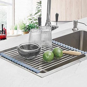 A roll-up dish rack with steel rods and a gray silicone edge rests over a kitchen sink, holding dishes and fruit.