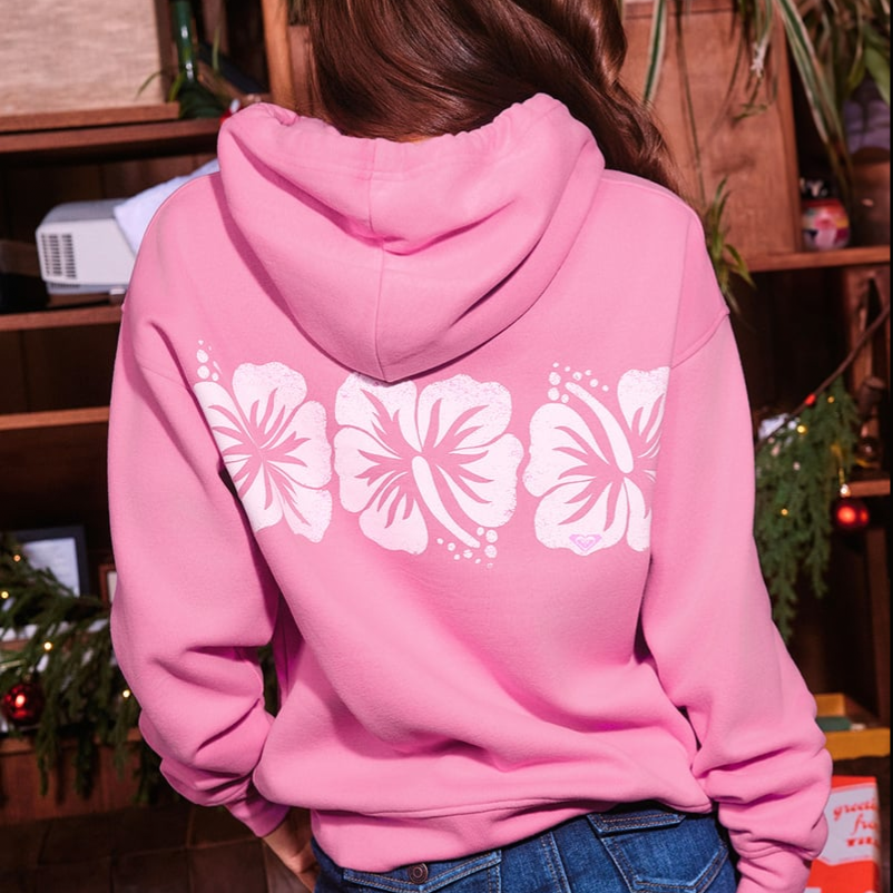 A person wearing a pink hoodie featuring a design of three white hibiscus flowers on the back.