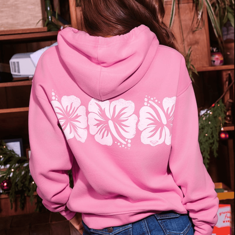 A person wearing a pink hoodie featuring a design of three white hibiscus flowers on the back.