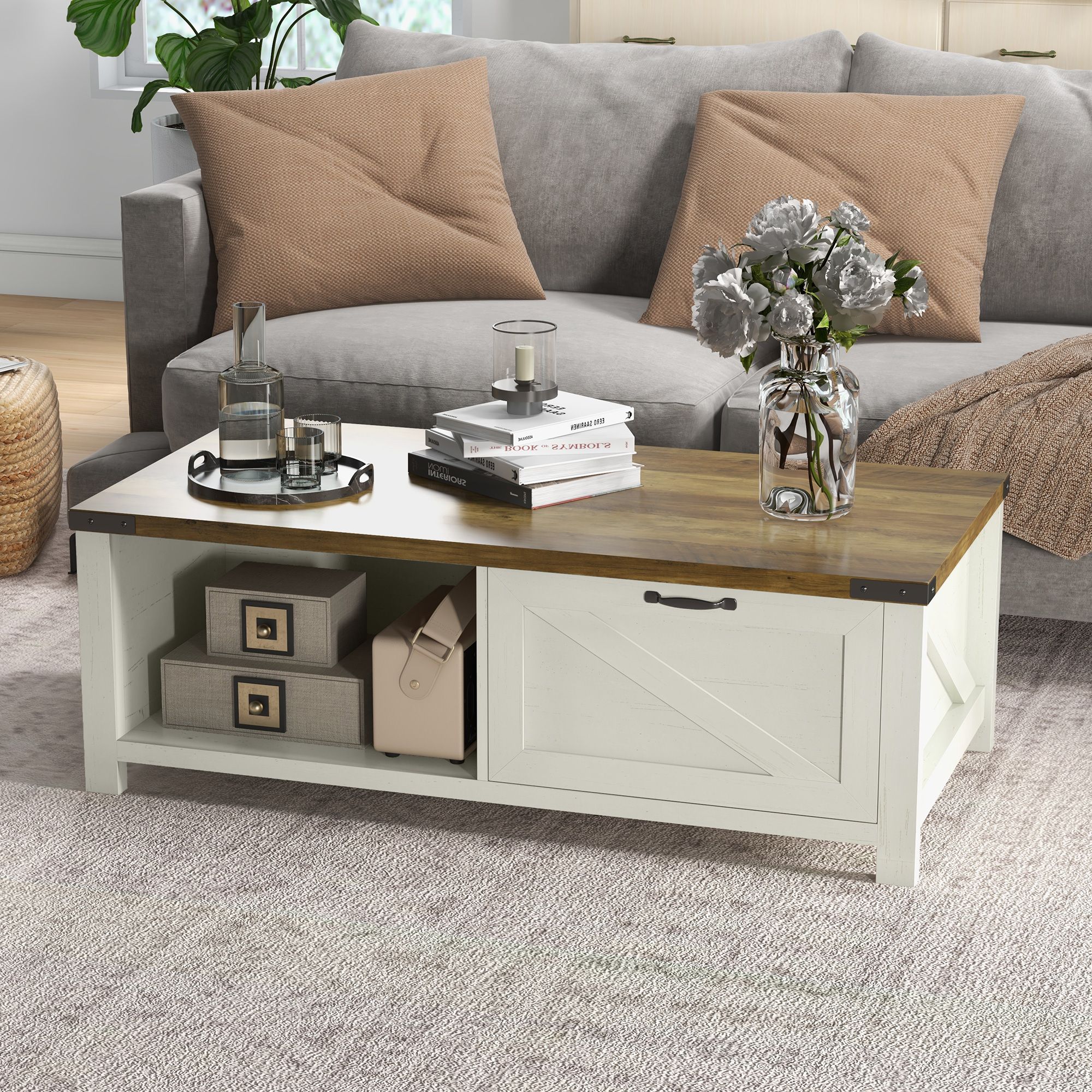 A rustic coffee table with a wooden top and white storage base featuring a sliding door. It is accessorized with books, a vase with flowers, a tray with glass items, and is placed in front of a gray sofa with beige pillows.
