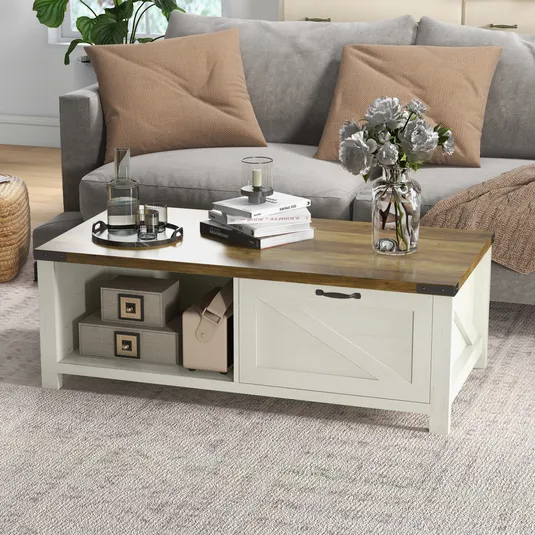 A rustic coffee table with a wooden top and white storage base featuring a sliding door. It is accessorized with books, a vase with flowers, a tray with glass items, and is placed in front of a gray sofa with beige pillows.