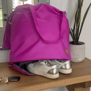A vibrant pink nylon tote bag is perched on a table, with a special compartment underneath that's partially unzipped revealing a pair of silver-colored shoes.