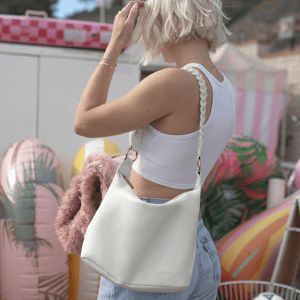 A person with short blond hair is wearing a white tank top and jeans, holding a white handbag with a braided strap.