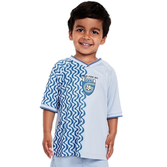 A young child wearing a light blue sports jersey with dark blue wave patterns along one side and the text \“GOING FOR MY GOALS\“ near a soccer-themed logo.