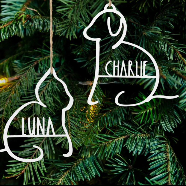 Two white metal ornaments in dog and cat shapes with names \“Charlie\“ and \“Luna\“ are hanging on a Christmas tree.