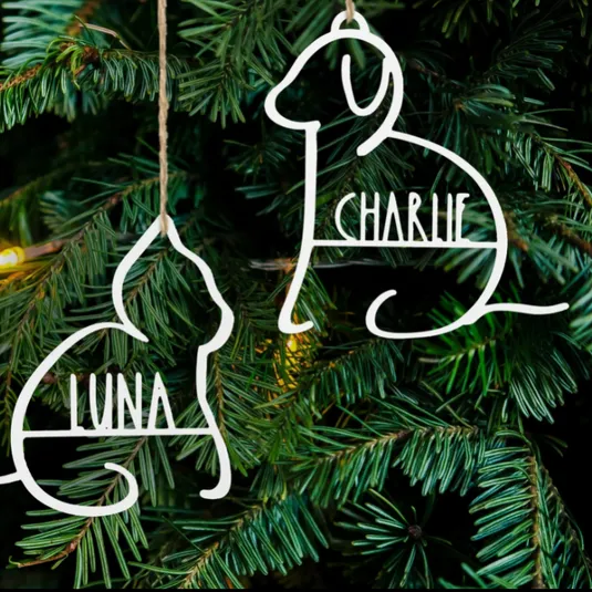 Two white metal ornaments in dog and cat shapes with names \“Charlie\“ and \“Luna\“ are hanging on a Christmas tree.