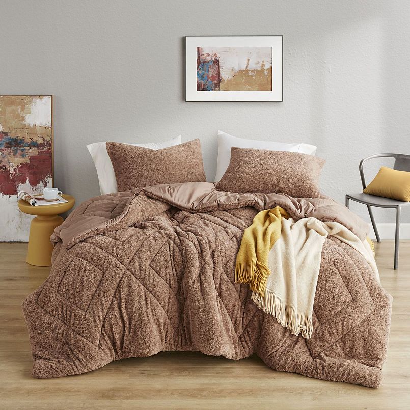 A cozy bedroom setup features a brown quilted comforter, matching pillows, and a draped yellow and cream fringe blanket. Accompanying decor includes abstract wall art, a round yellow side table, and a gray chair with a yellow cushion.