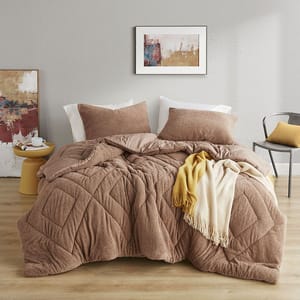 A cozy bedroom setup features a brown quilted comforter, matching pillows, and a draped yellow and cream fringe blanket. Accompanying decor includes abstract wall art, a round yellow side table, and a gray chair with a yellow cushion.
