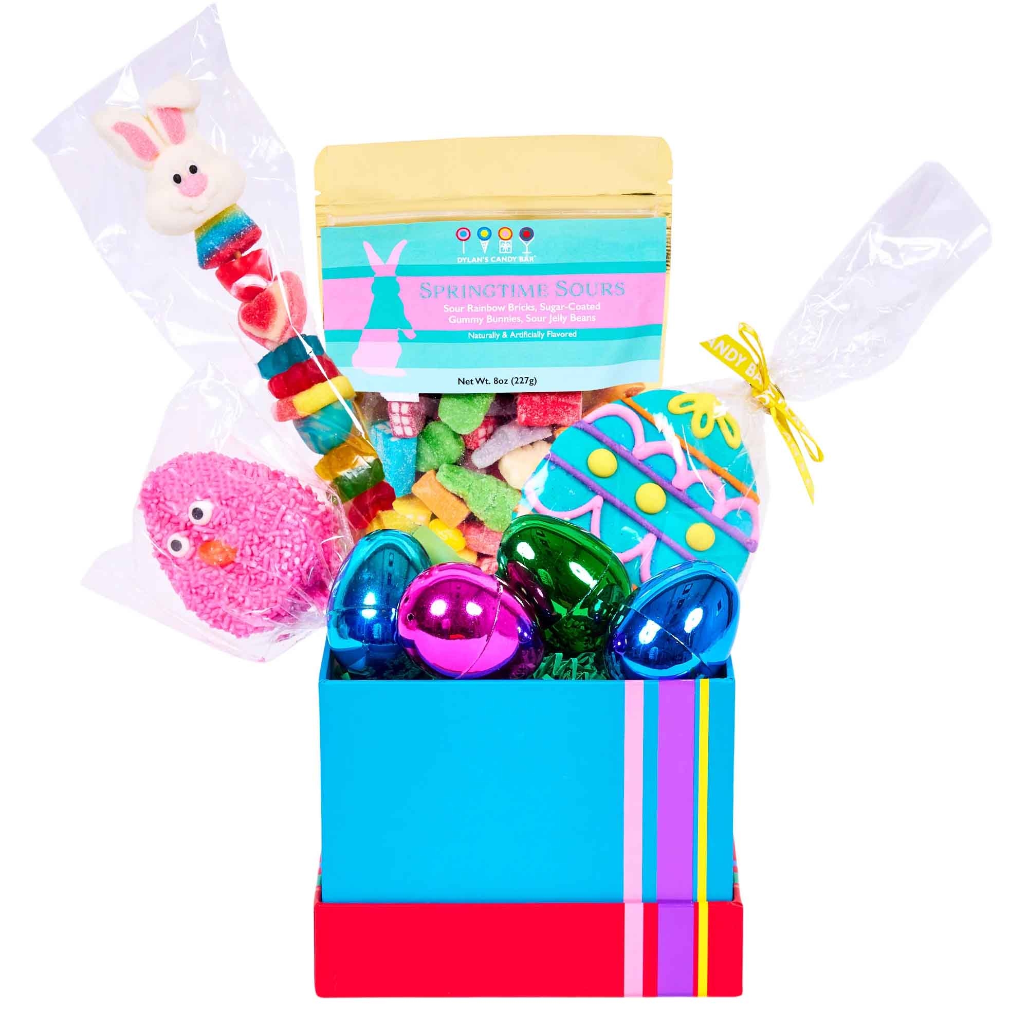 A festive gift basket featuring a colorful bunny-themed candy pop, a bag of \“Springtime Sours\“ gummy candies, a pink chick-shaped treat, a decorated egg-shaped cookie with icing, and shiny metallic eggs, all arranged in a vibrant blue and pink striped box.