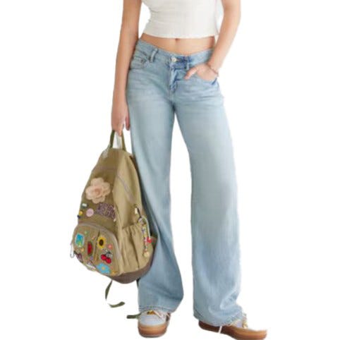 A person wearing light blue wide-leg jeans and a white crop top is holding a tan backpack decorated with colorful patches and designs.