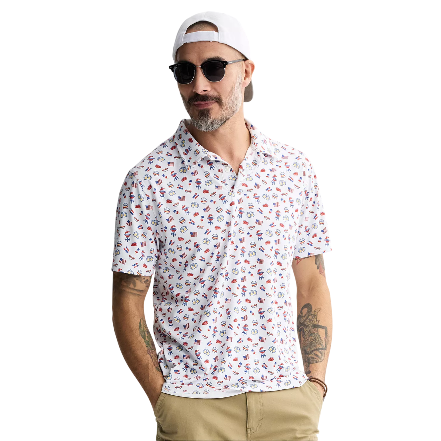 A man wearing a white polo shirt with quirky American-themed graphics, including flags and fireworks, paired with khaki shorts. He's accessorized with sunglasses, a backward white cap, and has tattoos on his left arm.