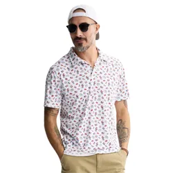 A man wearing a white polo shirt with quirky American-themed graphics, including flags and fireworks, paired with khaki shorts. He's accessorized with sunglasses, a backward white cap, and has tattoos on his left arm.