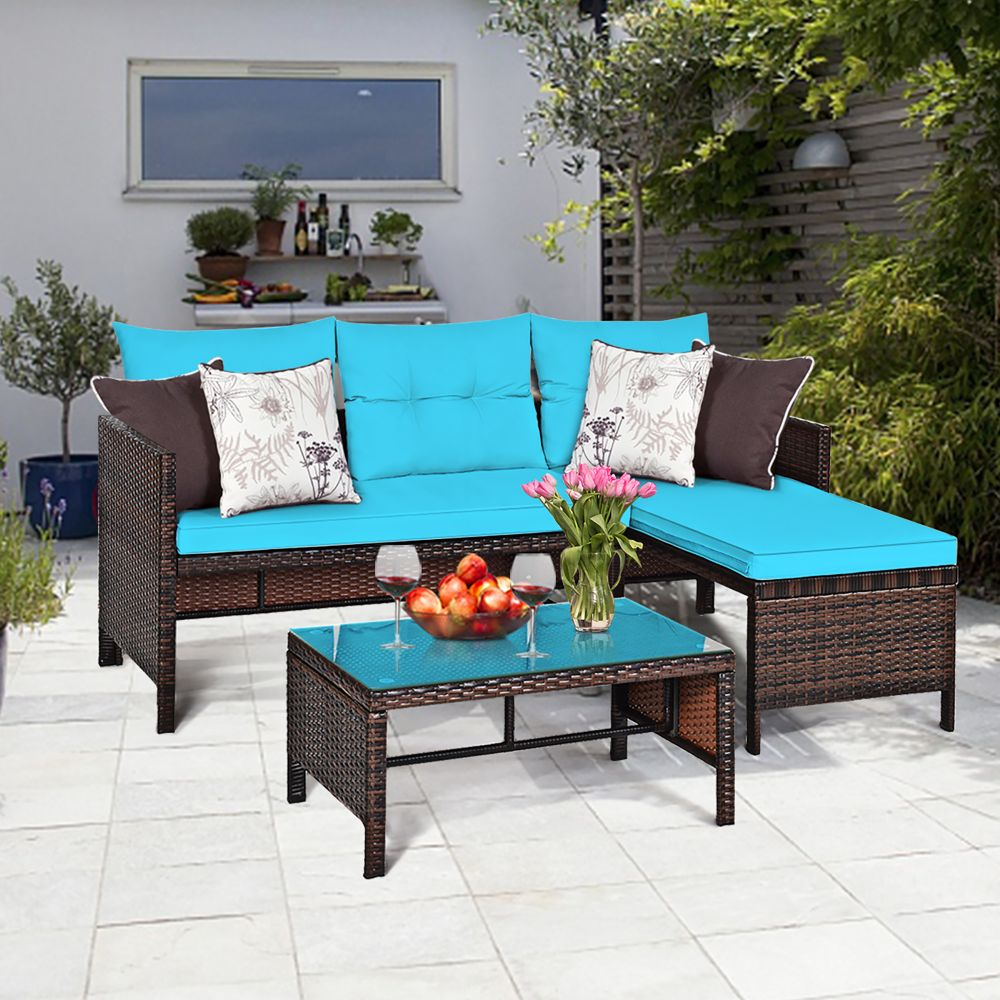 Outdoor patio set consisting of a wicker sectional sofa with blue cushions, a matching coffee table with a blue top, decorative pillows, and a vase with pink tulips, staged on a tiled patio with plants.