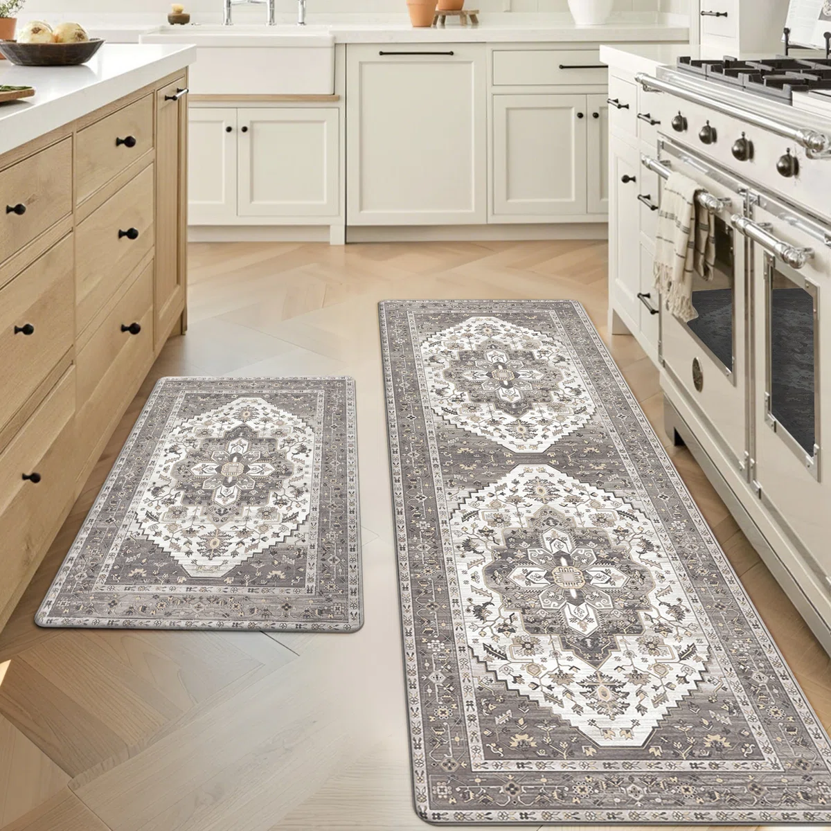 Kitchen features two decorative rugs with intricate patterns in a neutral color palette, placed on a light wood herringbone floor. One rug is a runner by the stove, and the other is a smaller mat by the sink.