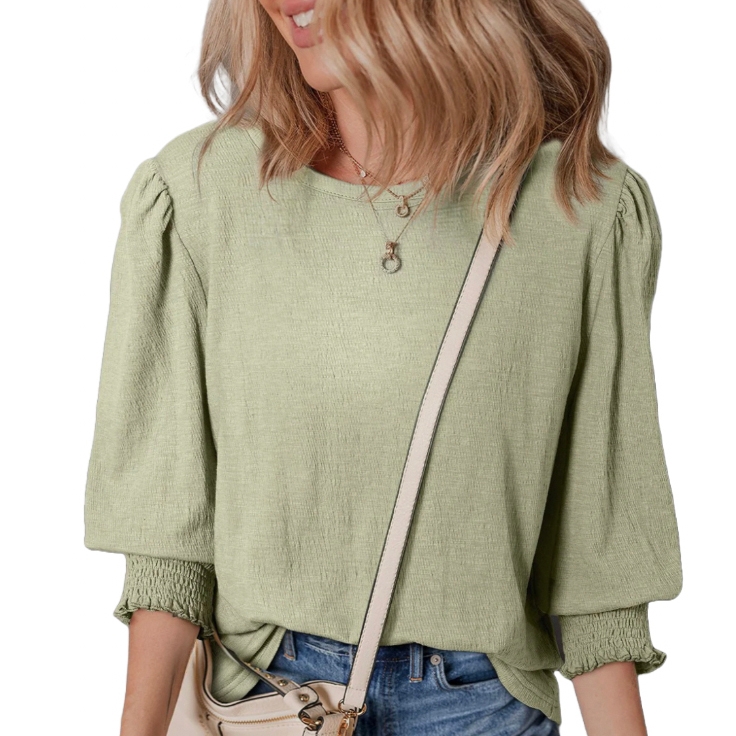 A light green, textured blouse with puffed three-quarter sleeves and elasticized cuffs, paired with blue jeans and a light-colored crossbody bag. The outfit is accessorized with a delicate necklace.