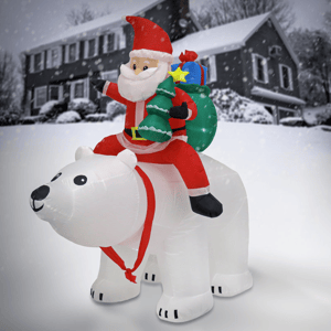 Inflatable Santa in a red suit rides a large white polar bear, holding gifts and a small Christmas tree, against a snowy house backdrop.