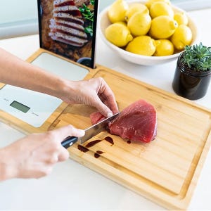 A person is slicing raw meat on a bamboo cutting board that integrates a digital scale and a phone holder, with a bowl of lemons and a small potted plant nearby.