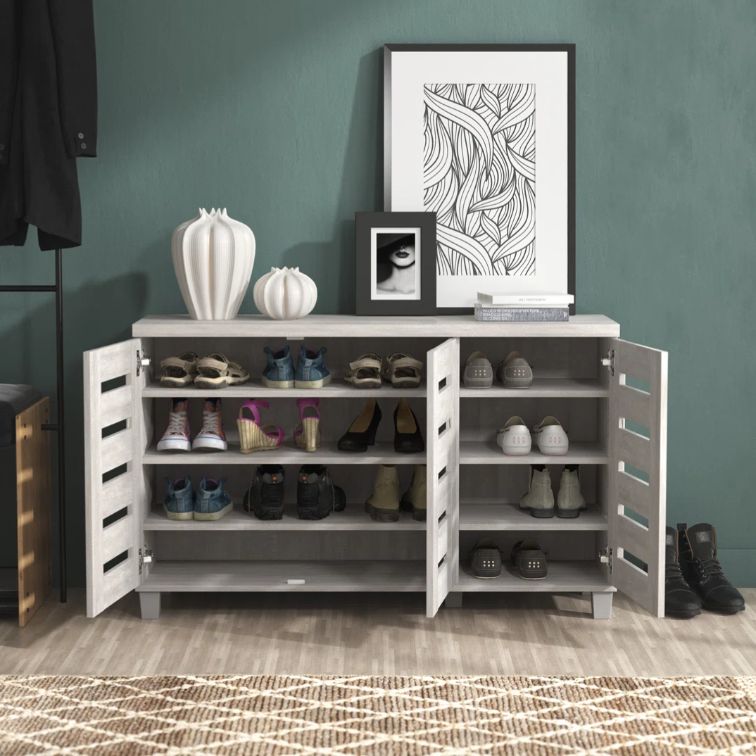 A modern shoe storage cabinet with two doors is shown, featuring multiple shelves holding various shoes. The top is decorated with white vases, framed artwork, and books, set against a textured teal wall.