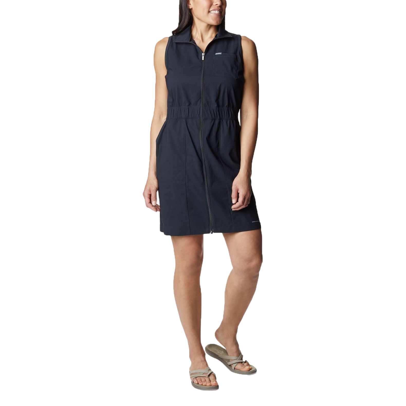 A woman is standing wearing a sleeveless black dress with a zippered front and sandals.