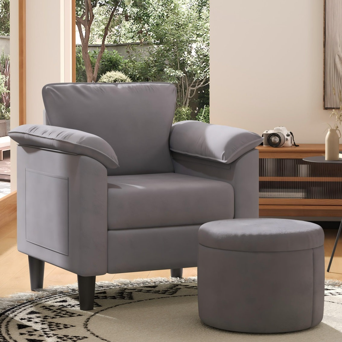 A gray armchair with padded arms and a matching round ottoman is placed on a patterned rug in a living room setting with natural light and greenery visible outside.