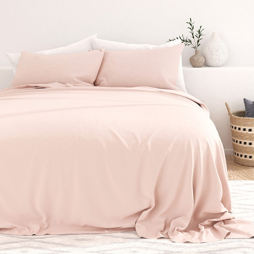 Soft pink bed set with two pillows on a white rug, accompanied by two textured vases and a basket with two cushions on a wooden floor.