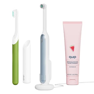 A green manual toothbrush, a blue and white electric toothbrush with charging stand, and a tube of Quip whitening anticavity fluoride toothpaste in watermelon flavor.
