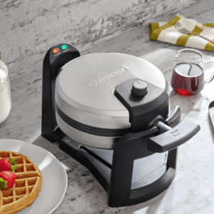 A waffle maker with a \“Cuisinart\“ branding is on a countertop, surrounded by a plate with waffle and strawberries, a jar of flour, syrup in a glass pitcher, and a dish with butter and a fork.