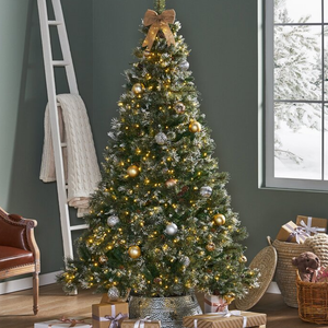 A 7-foot pre-lit Christmas tree with needle pine branches, featuring warm yellow lights, gold and silver ornaments, topped with a bow. It stands in a decorative pot surrounded by wrapped gifts and holiday decor.
