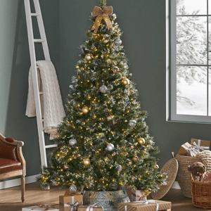 A 7-foot pre-lit Christmas tree with needle pine branches, featuring warm yellow lights, gold and silver ornaments, topped with a bow. It stands in a decorative pot surrounded by wrapped gifts and holiday decor.
