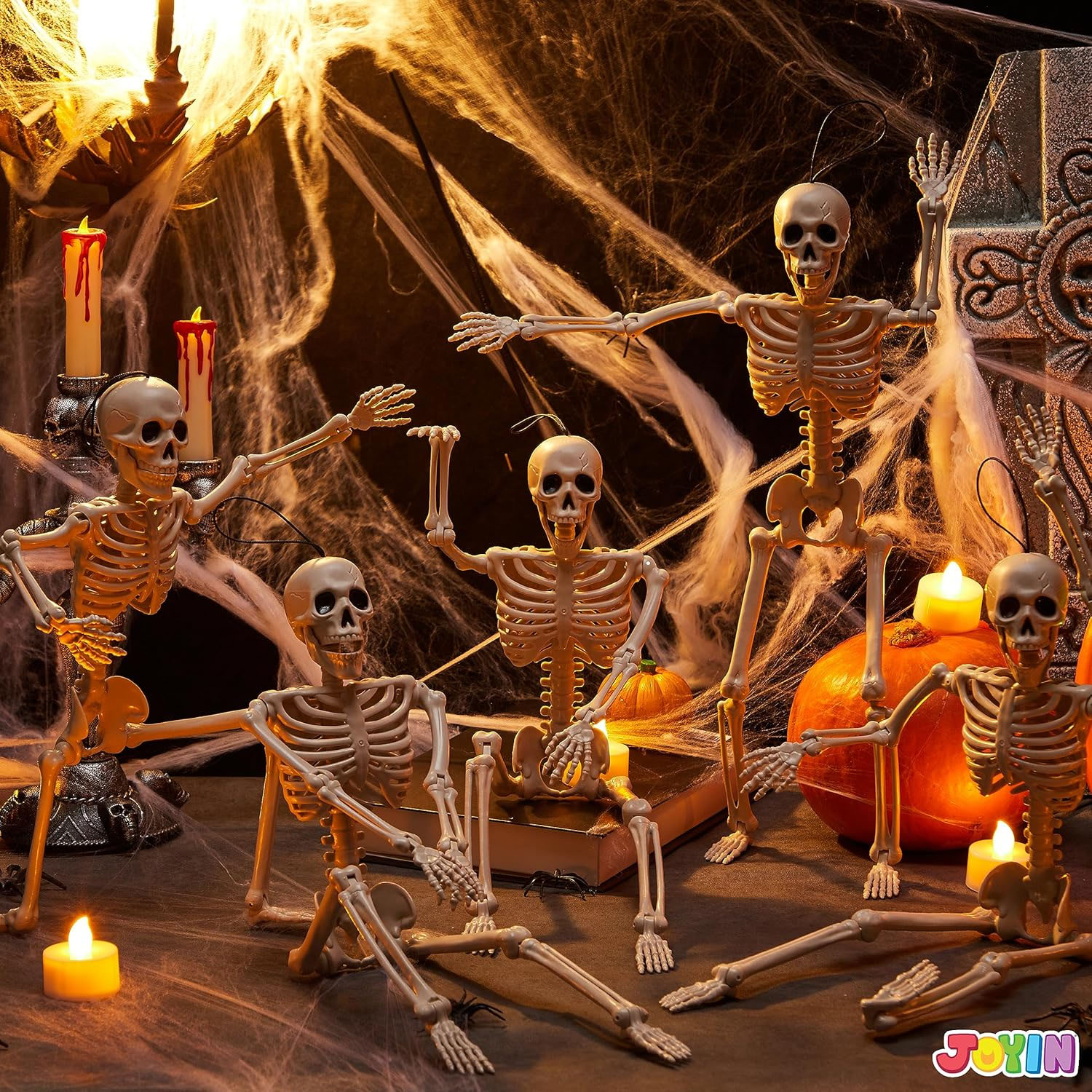 Five poseable skeleton figures are set in a spooky Halloween scene with cobwebs, candles, and pumpkins.
