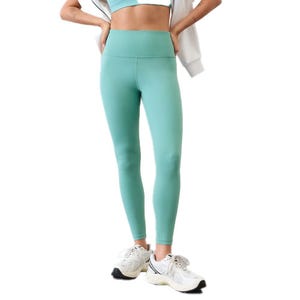 A woman wearing high-waisted, teal-colored leggings styled with a matching sports bra, white sneakers, and a white jacket draped over her shoulders.