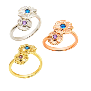 Three rings featuring double flower designs with birthstones: silver with a blue and a purple gemstone, rose gold with a blue and a purple gemstone, and gold with a blue and a purple gemstone.
