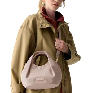 A person in a tan jacket and pink collared shirt holds a light pink handbag with a round handle and shoulder strap.
