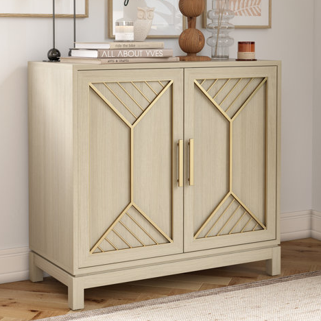 A compact storage cabinet with light wood finish and geometric gold metal accents on the doors, featuring two handles and elevated on short legs, suitable for modern decor.
