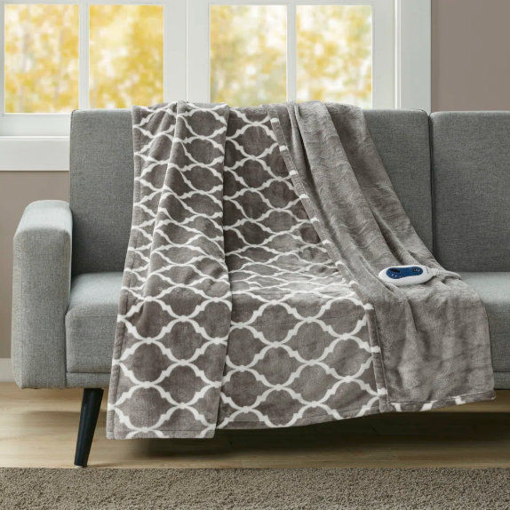 A gray sofa is adorned with a patterned gray and white electric blanket featuring a remote control. A side table holds a potted plant and a lamp stands nearby, set against a backdrop of large windows with an autumn view.